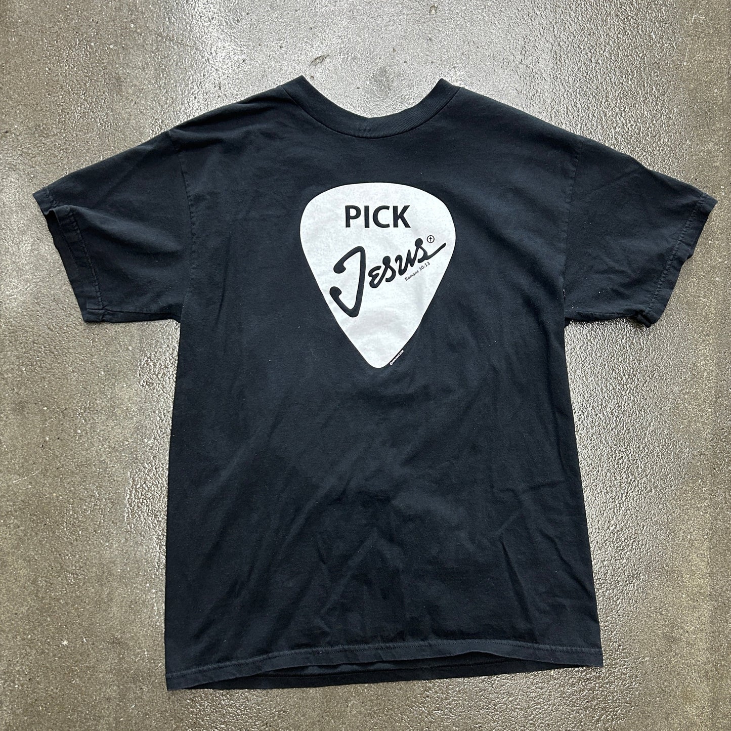Black t-shirt with a white guitar pick graphic and 'Pick Jesus' text on a gray surface