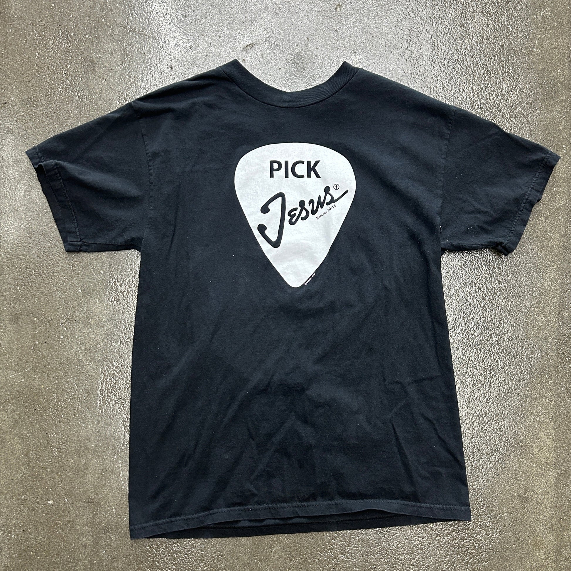 Black t-shirt with a white guitar pick graphic and 'Pick Jesus' text on a gray surface